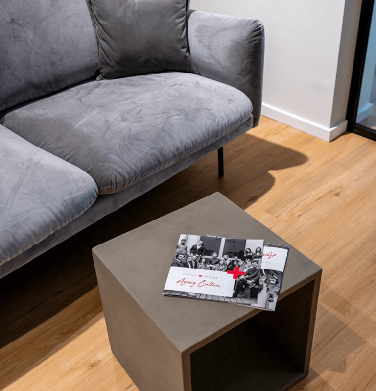 Auf dem Holzboden steht ein graues Sofa neben einem quadratischen grauen Couchtisch. Darauf liegen zwei Broschüren mit der Aufschrift „Digitalspital“ – Schwarzweißfotos mit auffälligem roten Text –, die dem ruhigen Raum einen modernen Touch verleihen.