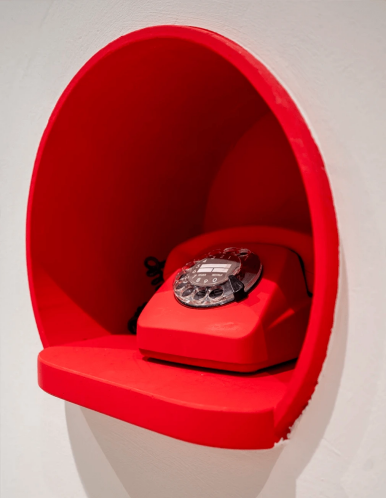 Ein rotes Vintage-Wählscheibentelefon steht in einer runden roten Nische vor einer weißen Wand und verbindet nostalgischen Charme mit dem digitalen Zeitalter. Die Wählscheibe ist sichtbar, und unter der Nische befindet sich ein kleines Regal.