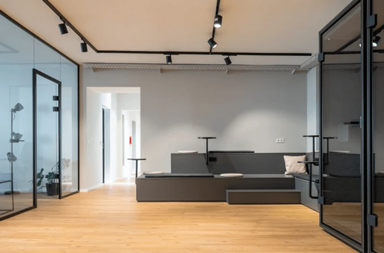 Moderne Büroräume mit digitalen Krankenhauselementen, darunter Konferenzräume mit Glaswänden, Holzböden und graue Sitzbereiche. Schienenbeleuchtung ziert die Decke und unterstreicht das minimalistische Design mit einer neutralen Farbpalette.