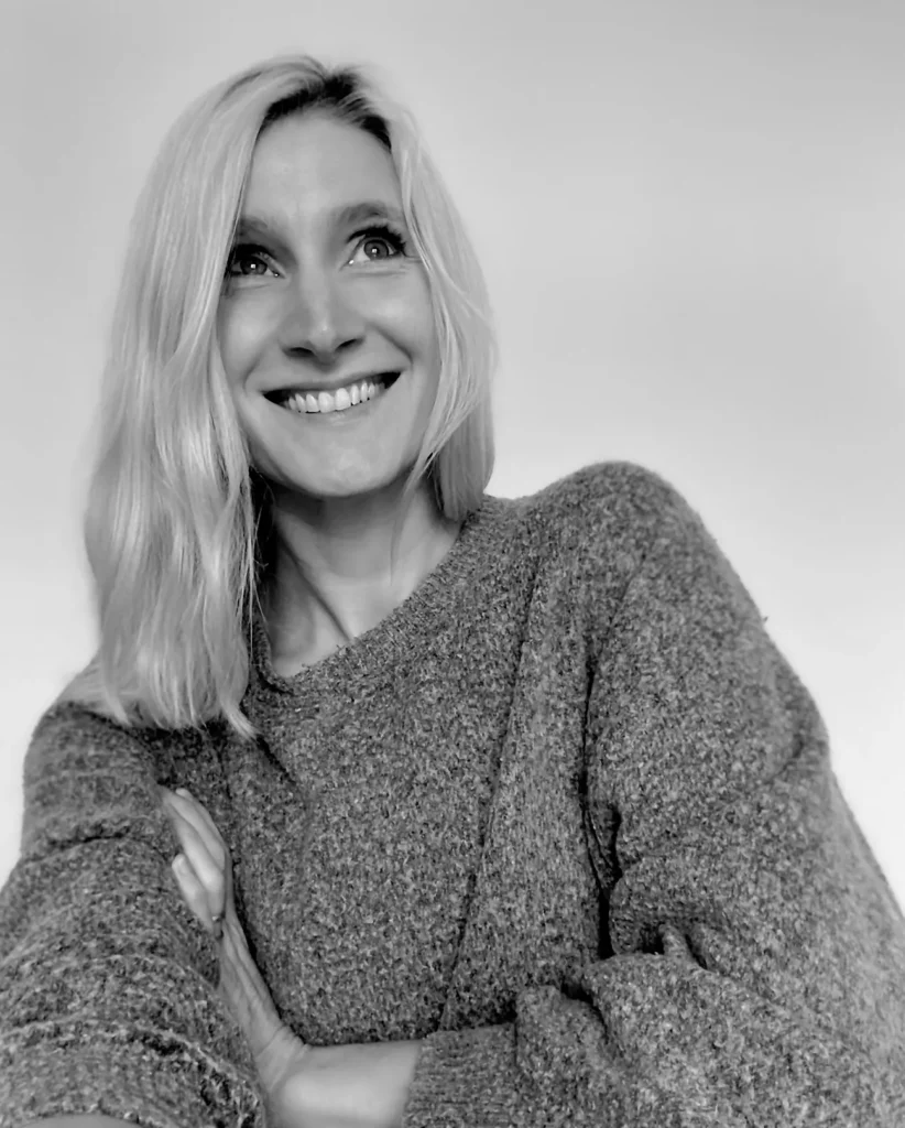 Schwarz-Weiß-Bild einer Person mit schulterlangem blondem Haar, die einen kuscheligen Strickpullover trägt, lächelt und vor einem schlichten Hintergrund leicht zur Seite blickt. Ihre Arme sind in entspannter Pose verschränkt und halten in diesem digitalen Krankenhausporträt einen zeitlosen Moment fest.