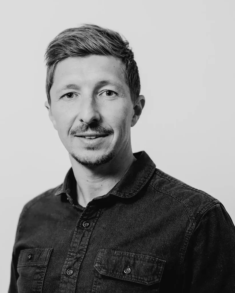 Ein Schwarz-Weiß-Porträt von Digitalspital zeigt eine Person mit kurzen Haaren und dezentem Bartwuchs, die ein Hemd mit Knöpfen trägt. Ihr Blick trifft vor einem schlichten Hintergrund direkt auf die Kamera und verkörpert eine Mischung aus Modernität und zeitloser Schlichtheit.
