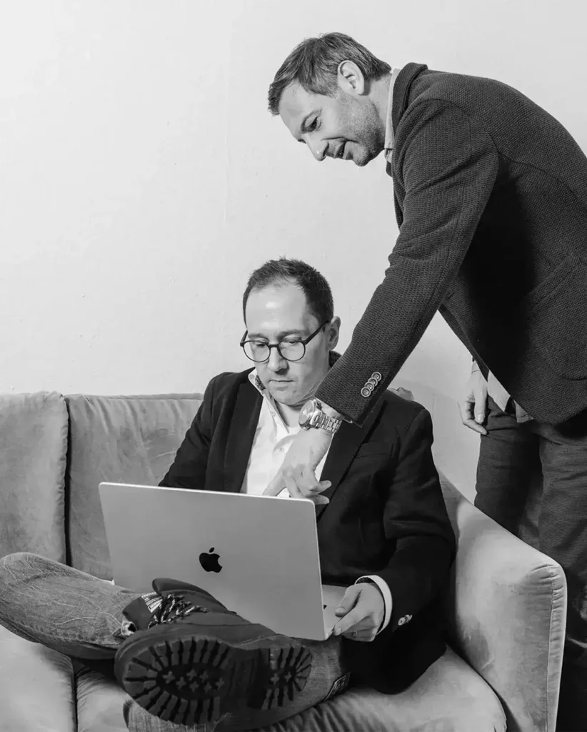Auf einem Schwarzweißfoto sind zwei Männer zu sehen. Einer sitzt mit einem Laptop auf einem Sofa, der andere steht vorgebeugt und zeigt auf den Bildschirm. Beide tragen Anzüge. Die Szene spielt in einem schlichten Raum mit einer weißen Wand.