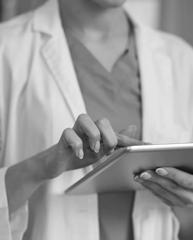 Eine Person im Laborkittel benutzt ein Tablet und tippt mit den Fingern auf den Bildschirm. Der Fokus liegt auf ihren Händen und dem Tablet, der Hintergrund ist unscharf. Sie scheint sich in einem professionellen oder klinischen Umfeld zu befinden.