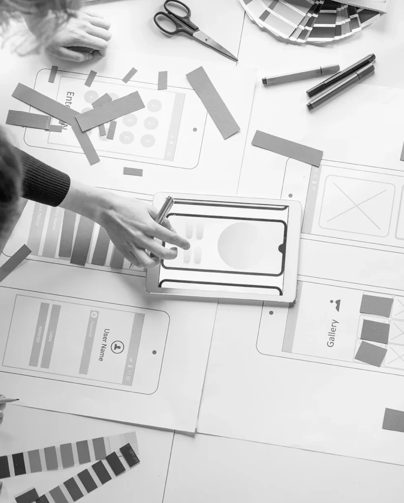 Ein Schwarz-Weiß-Bild zeigt Menschen, die an der Entwicklung einer mobilen App arbeiten. Auf einem Schreibtisch liegen Papierausschnitte von mobilen Bildschirmen mit Schaltflächen und Layouts. Scheren, Lineale, Stifte und Farbmuster liegen verstreut herum. Eine Hand richtet ein Papierelement.