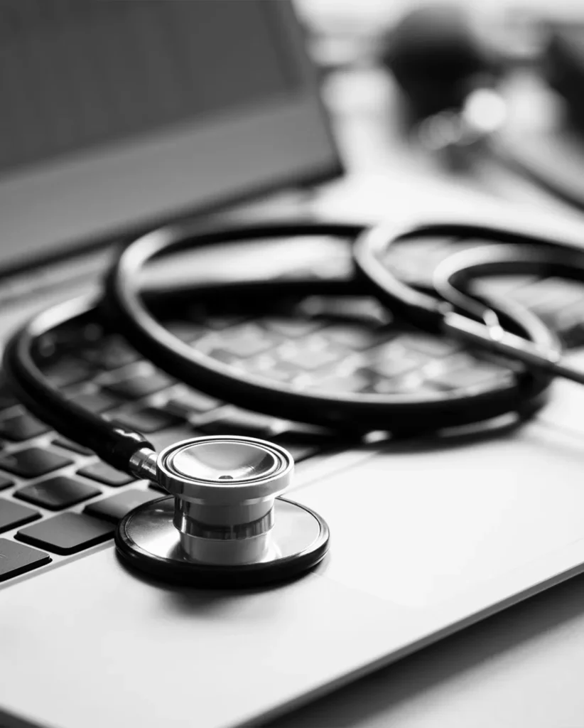 Ein Schwarz-Weiß-Bild zeigt ein Stethoskop auf einer Laptop-Tastatur. Der Fokus liegt auf dem Stethoskop, das die Schnittstelle zwischen Gesundheitswesen und Technologie symbolisiert. Der Hintergrund ist leicht unscharf.