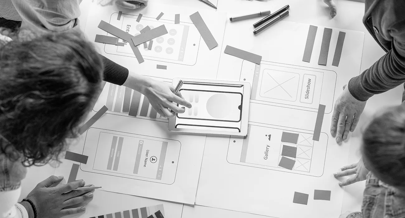 Menschen arbeiten gemeinsam an einem Tisch mit Wireframe-Designs für mobile Apps. Hände platzieren ausgeschnittene Elemente auf den Papieren, was auf ein Brainstorming oder eine Design-Sitzung hindeutet.