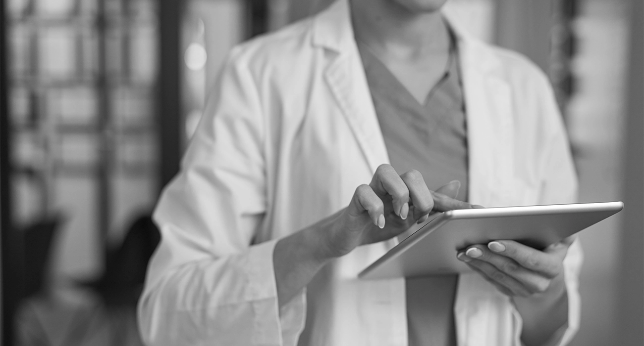 Eine Person in Laborkittel und OP-Kleidung bedient ein Tablet. Der Hintergrund ist unscharf, wodurch der Fokus auf der Tablet-Interaktion liegt. Schwarz-Weiß-Bild.