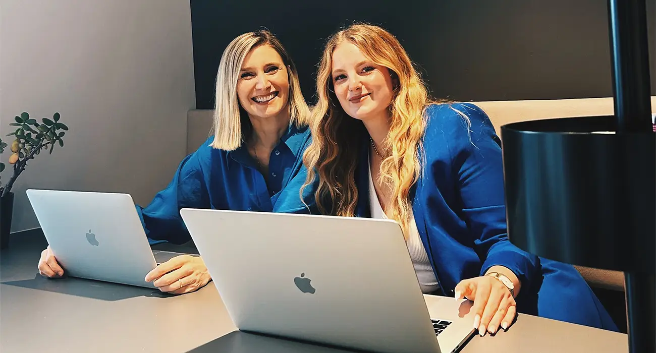 Zwei lächelnde Frauen in blauen Outfits sitzen an einem Tisch mit Laptops vor sich und verkörpern den Geist des Digitalspitals. Sie befinden sich in einer modernen Büroumgebung mit einer schwarzen Lampe und einer kleinen Pflanze auf dem Tisch.