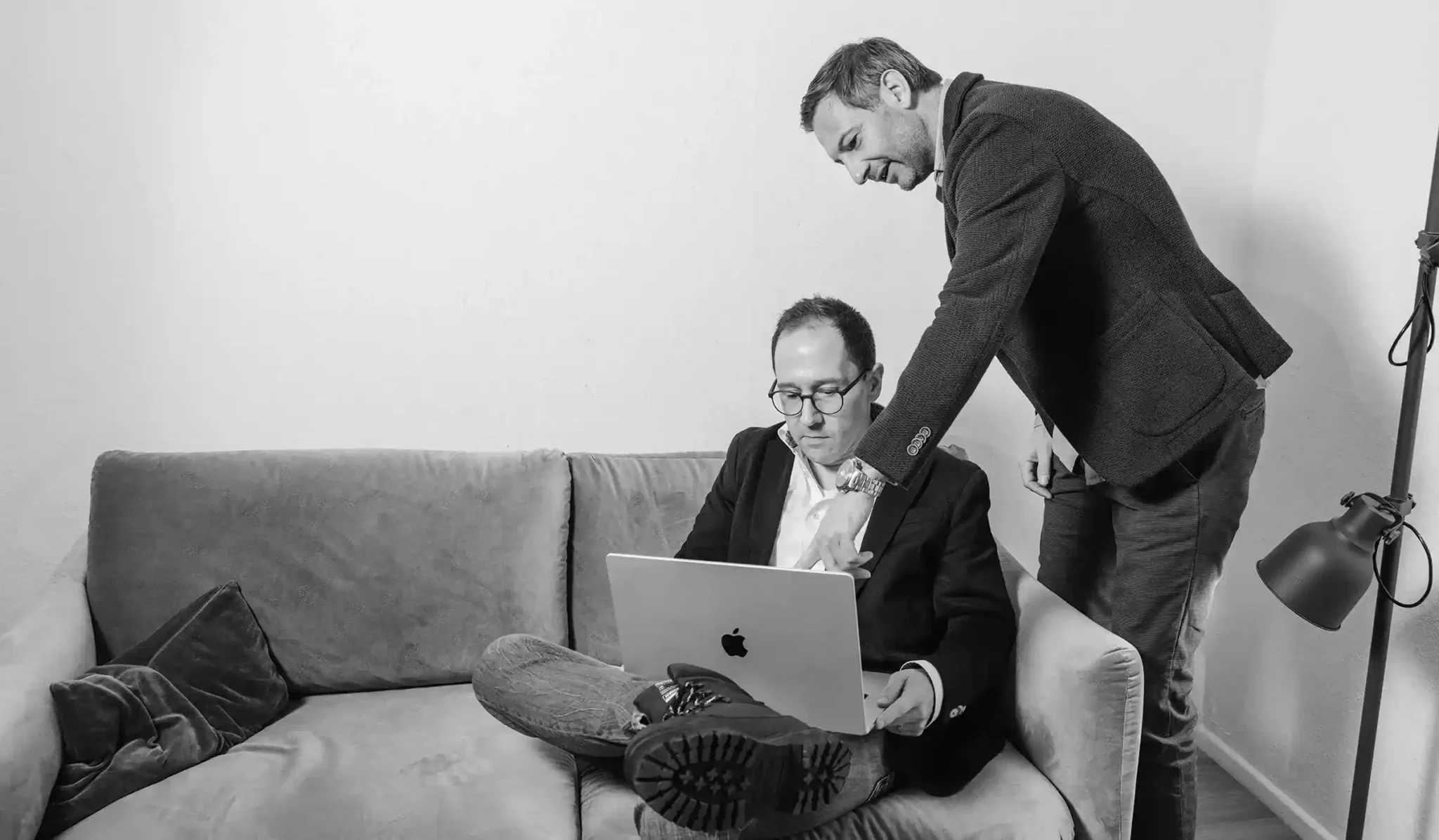 Zwei Männer in einem Raum mit einer Couch. Einer sitzt und benutzt einen Laptop, der andere steht daneben und zeigt auf den Bildschirm. Sie diskutieren. Die Atmosphäre wirkt professionell. Schwarz-Weiß-Foto.