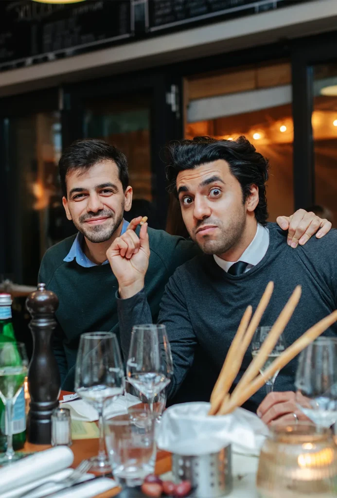 Zwei Männer sitzen an einem Restauranttisch. Vor ihnen stehen Teller und Gläser. Ein Mann lächelt und hat den Arm um den anderen gelegt. Der andere hält ein Grissini in der Hand und schneidet ein verspieltes Gesicht. Auf dem Tisch stehen Flaschen und eine Pfeffermühle.