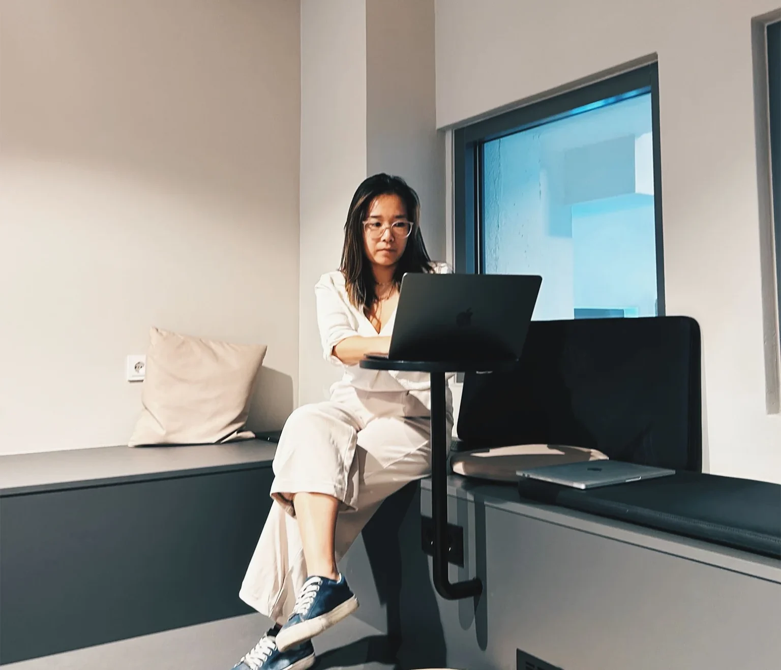 Eine Person sitzt auf einer Bank mit Kissen und benutzt einen Laptop auf einem kleinen Tisch. Sie trägt eine Brille, ein helles Outfit und Turnschuhe. Rechts ist ein Fenster mit teilweisem Blick nach draußen zu sehen.