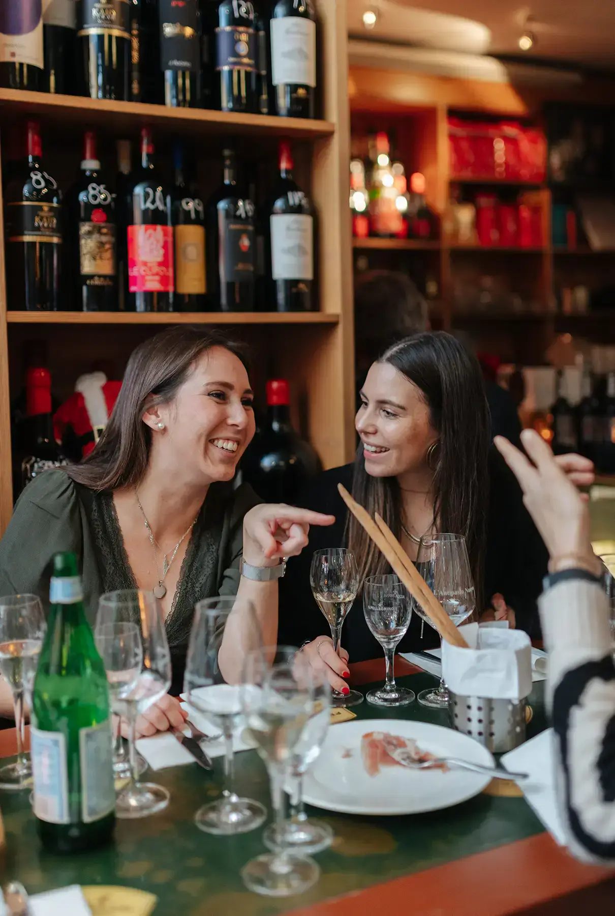 Zwei Frauen sitzen lächelnd an einem Tisch in einem Restaurant und unterhalten sich wie Kolleginnen, die gerade ihr neuestes Gesundheitsprojekt planen. Im Hintergrund stehen Weinflaschen in den Regalen. Gläser, eine Wasserflasche und Teller stapeln sich auf dem Tisch, während die beiden digitalen Impulsgeberinnen Ideen austauschen.
