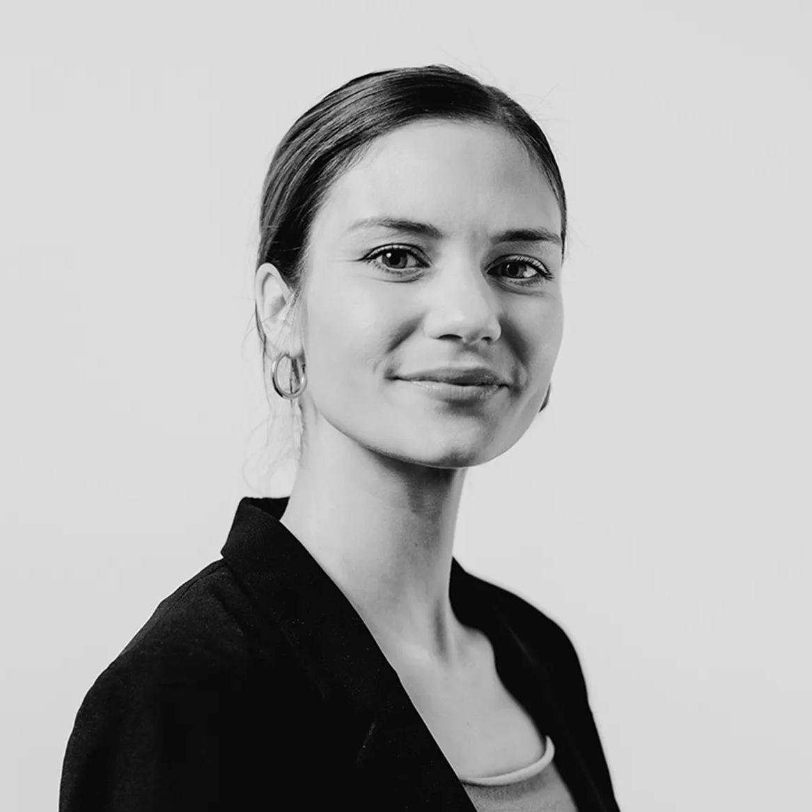 Schwarz-Weiß-Porträt einer Frau mit zurückgebundenen dunklen Haaren, die Reifohrringe, einen dunklen Blazer und ein helles Oberteil trägt und vor einem schlichten Hintergrund mit selbstbewusstem Ausdruck leicht zur Seite schaut.