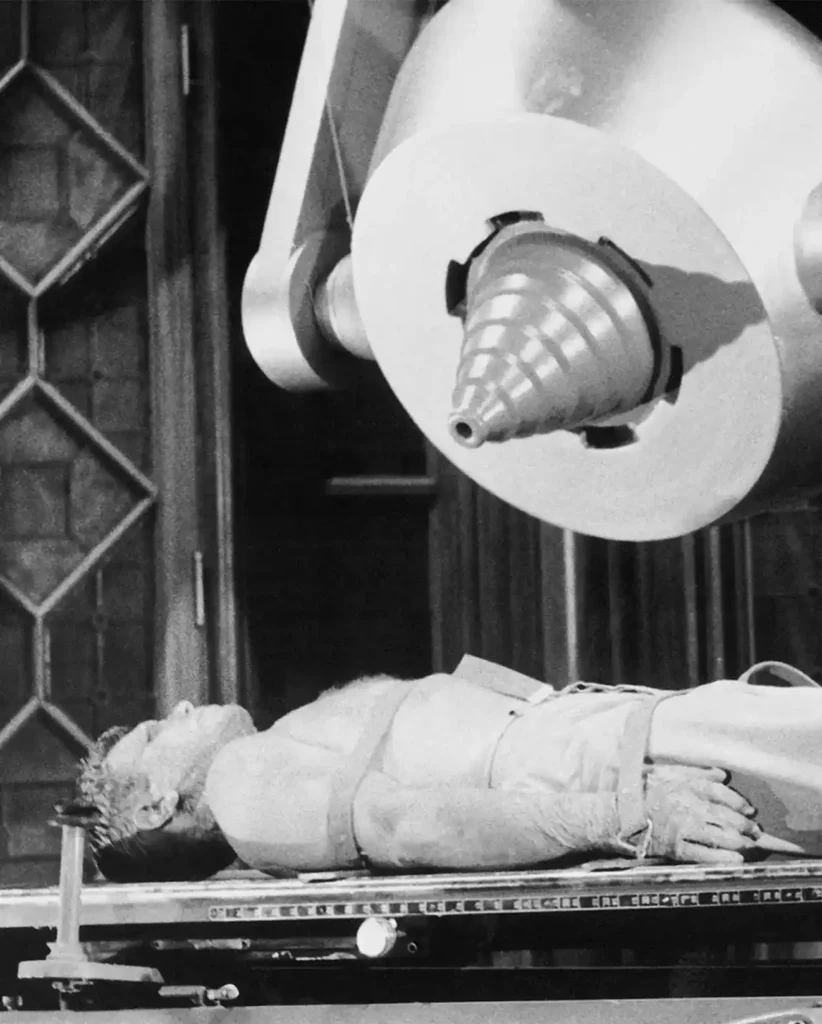 Ein Mann liegt auf einem Tisch unter einer großen, futuristischen, kegelförmigen Maschine in einer schwarz-weißen Vintage-Kulisse. Er scheint oberkörperfrei und trägt eine Hose. An seinen Armen trägt er Geräte oder Riemen. Die Szene erinnert an ein Retro-Science-Fiction- oder medizinisches Experimentalthema.