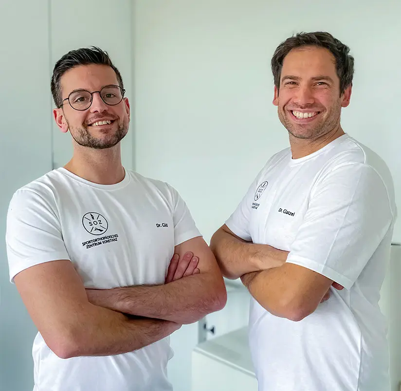 Zwei Männer in weißen Praxis-Marketing-T-Shirts mit verschränkten Armen stehen lächelnd nebeneinander in einem hellen Raum. Ihre Shirts zeigen ein kreisförmiges Logo und einen Text auf der linken Brust. Beide haben kurze Haare und eine Brille.